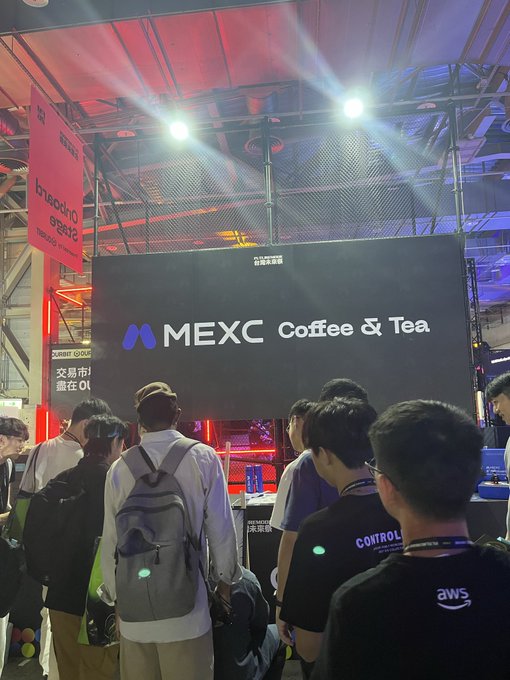 People stand in a queue at a busy convention booth with red lighting, facing a large screen displaying 'MEXC Coffee & Tea' and the KuCoin logo.