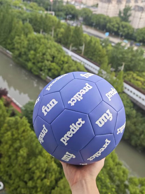 A hand holds a blue soccer ball branded with "predict" and "my" logos against a backdrop of a river and tree-lined walkway, visually promoting the prediction market platform "predict.fun" referenced in the post about Polymarket's deleted airdrop teaser.