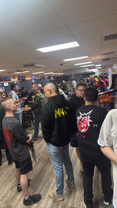 A crowded indoor venue, possibly a gaming or sports bar, filled with people standing and mingling under fluorescent lighting, with some wearing casual clothing like a black jacket with a red logo.