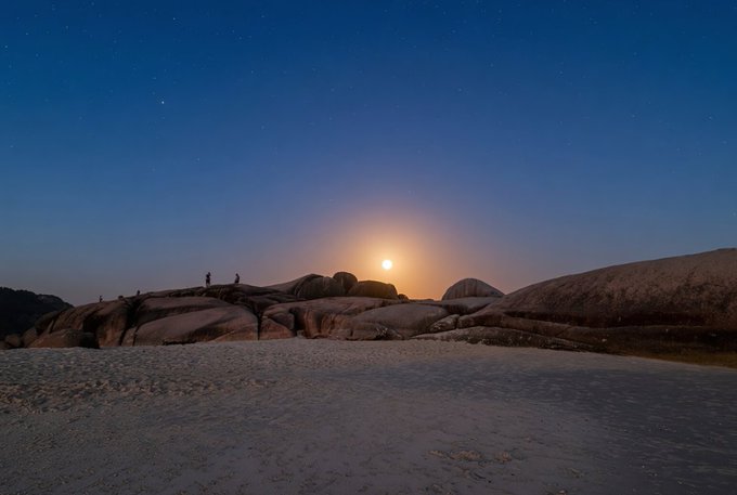 A serene night landscape featuring a full moon rising over a rocky beach with silhouetted figures, under a starry blue sky.