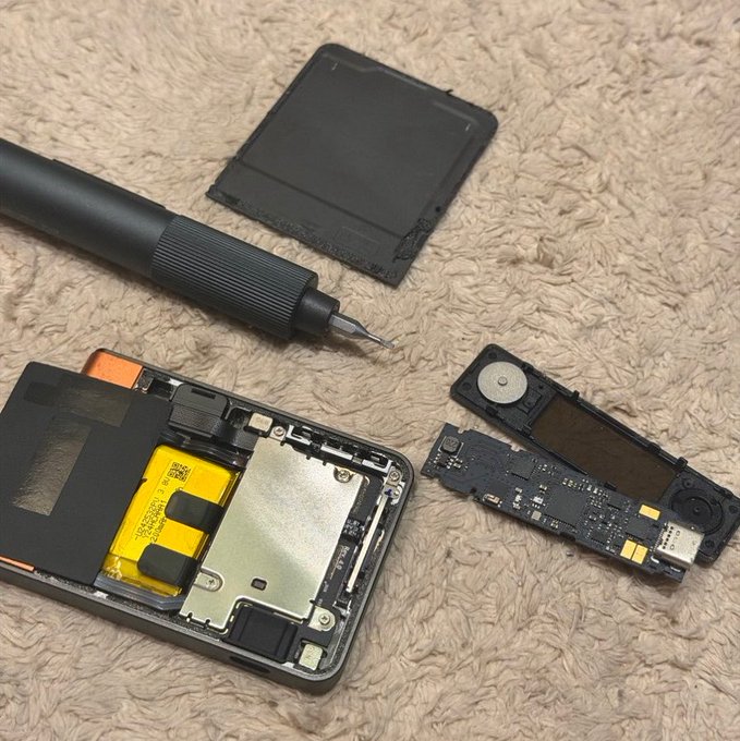 A disassembled smartphone with its back cover, circuit board, and battery exposed lies on a towel alongside a precision screwdriver, suggesting a repair in progress.