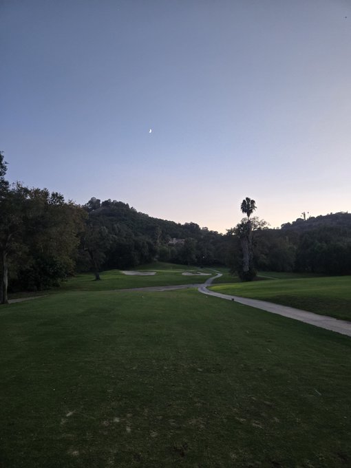 A scenic view of a golf course fairway at dusk with sand traps, palm trees, and a crescent moon visible in the sky.