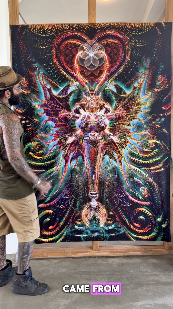 A heavily tattooed man in a hat stands next to a large, colorful psychedelic tapestry featuring symmetrical imagery and a heart, with text reading 'CAME FROM' at the bottom.