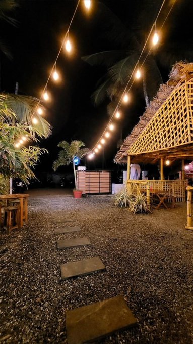 A nighttime beachside scene with string lights, a thatched-roof bamboo hut, wooden tables, and stepping stones across pebbled ground.
