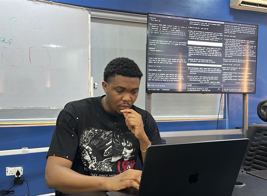 A young man in a black graphic t-shirt sits at a laptop (Apple MacBook) in front of a screen displaying terminal/command-line code, with his hand thoughtfully on his chin as he works. Behind him is a whiteboard with red writing and a blue wall, while the screen shows developer commands referencing Sui blockchain network work, matching the post about building AI integrations and partnerships with Sui Network protocols like DeepBook, Hudi, and Walrus.