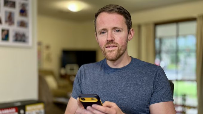 A man in a gray t-shirt holds a smartphone while speaking, looking directly at the camera in a home setting. The image suggests him engaging with AI agents or blockchain technology, visually supporting the post's discussion about BAP-578 standards and BORT agents on BNB Chain.