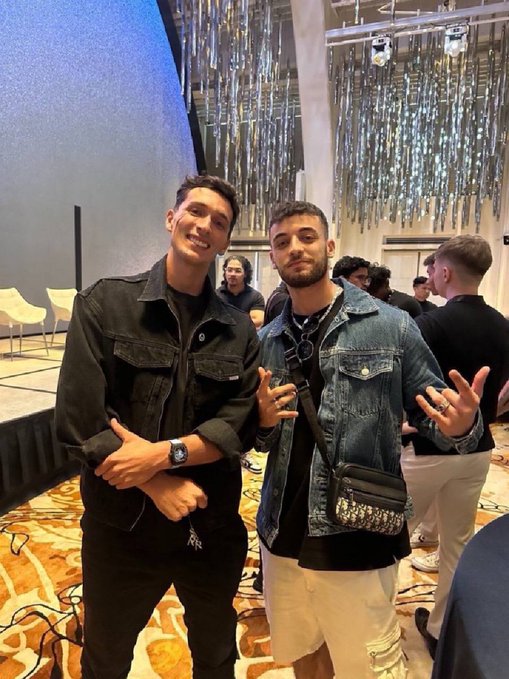 Two men posing together indoors, one in a black denim jacket and the other in a blue denim jacket with a crossbody bag, set against a backdrop of silver streamers.