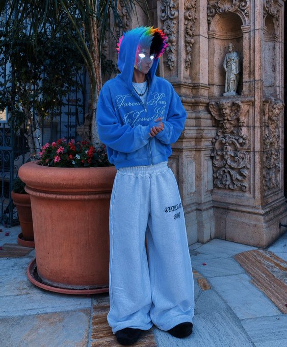 A person wearing a blue hoodie and oversized light blue pants stands in front of an ornate stone building, featuring a digital rainbow-colored hair filter effect.