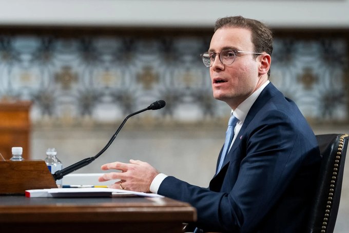 A man in a navy suit, glasses, and blue tie speaks into a microphone while seated at a wooden desk, with water bottles and papers in front of him.