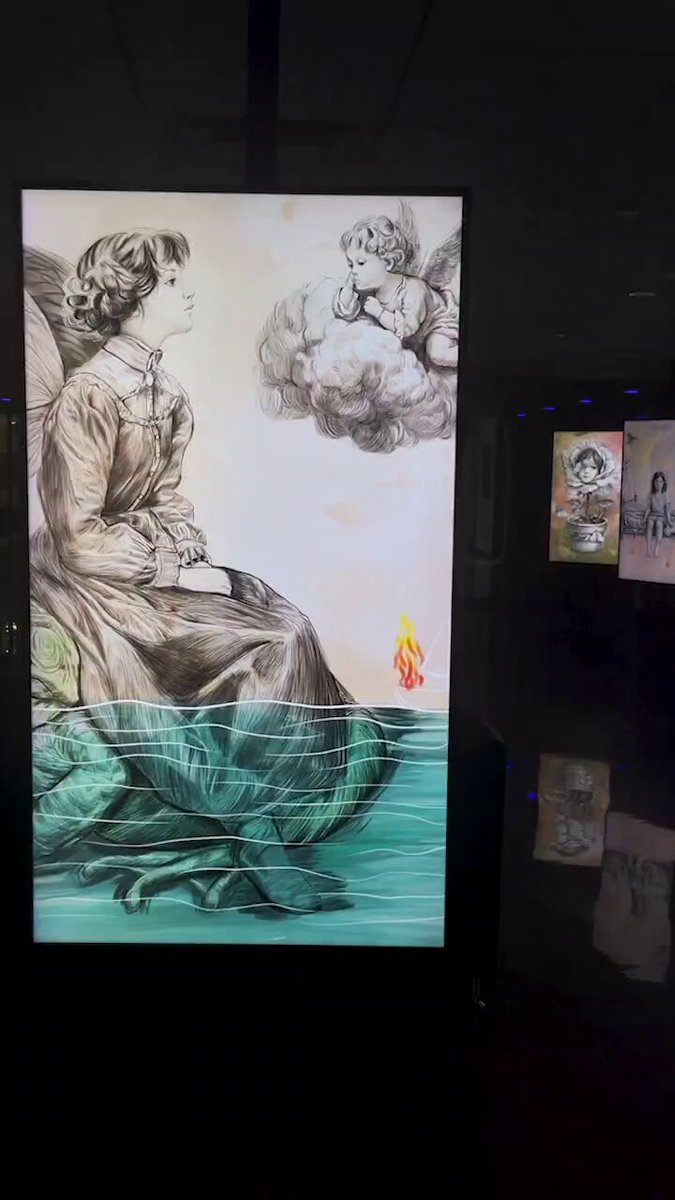The image features a painting of an angel sitting on a rock, with a fire burning below her feet. The painting is displayed against a black background, which contrasts with the white wall behind it. The artist's signature can be seen at the bottom right corner of the painting.