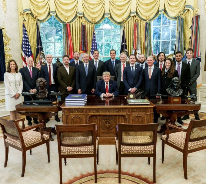 A formal group photo in the Oval Office at the White House showing Trump seated at the Resolute Desk surrounded by administration officials and cabinet members, with U.S. flags visible behind them.