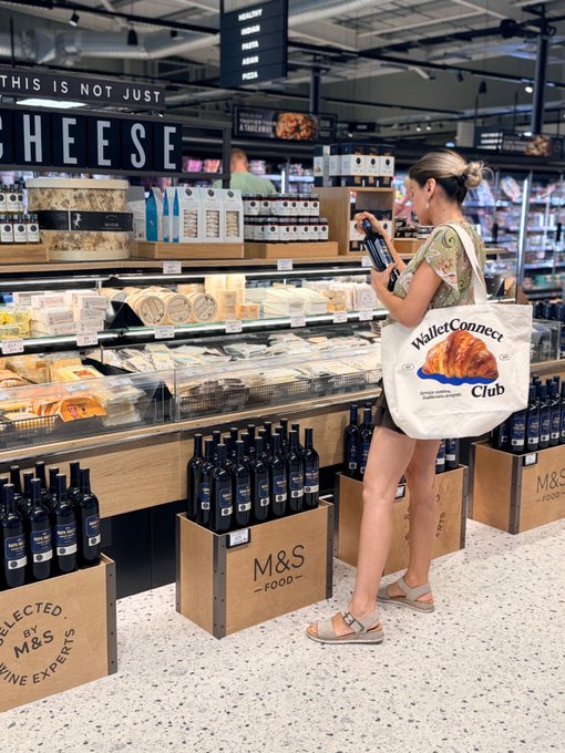 A woman with a 'WalletConnect Club' tote bag browses the cheese section in an M&S Food store, holding a bottle while standing near crates of wine.
