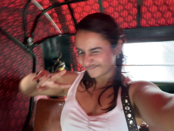 A casual, slightly blurry selfie of a young woman smiling and winking at the camera, wearing a pink top with decorative straps, against a backdrop of red padded upholstery.
