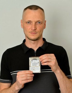 A man in a black polo shirt holding up a small retail hard drive package.