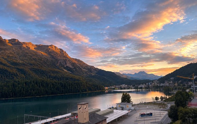 A scenic landscape view of a calm lake reflecting a dramatic sunset sky, surrounded by mountains and a small town along the shore.