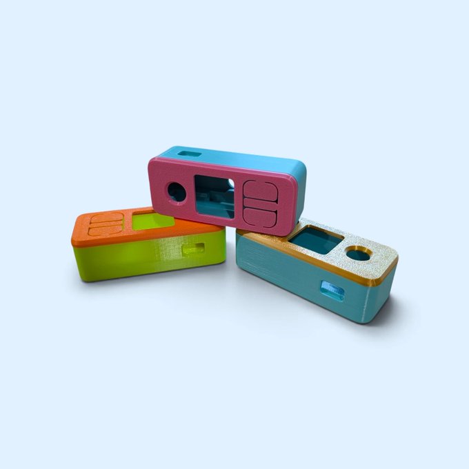 Three colorful 3D-printed rectangular camera-like devices stacked on a light blue background, featuring pink, green, orange, and teal colors with various openings and details.