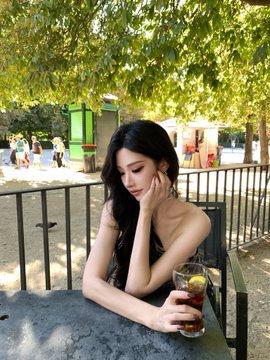 A portrait-style photo of a young woman with long dark hair sitting at an outdoor table under trees, holding a glass of iced tea.
