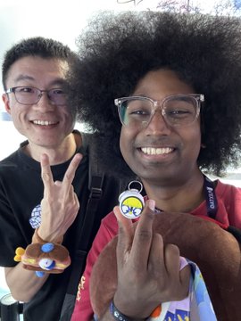 Two smiling young people pose closely together for a photo, each making finger hearts; one wears glasses and a black shirt with a cartoon keychain, the other has curly hair, clear glasses, and a red shirt.