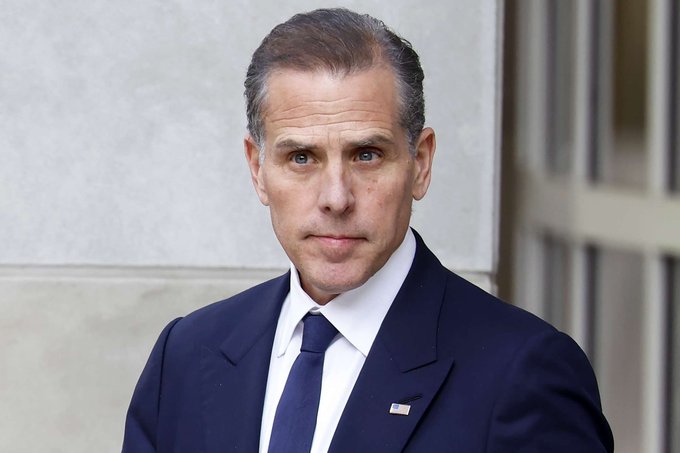 A man in a navy suit with a white shirt and dark tie, wearing an American flag lapel pin, standing outdoors against a light stone background.