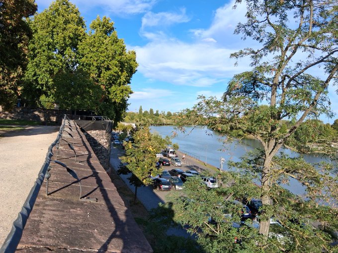 A scenic view from an elevated walkway showing a river, a parking area with several cars, and lush green trees under a blue sky.