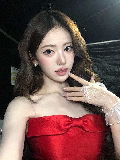 A young woman with long brown hair poses wearing a red strapless top and white lace fingerless glove, with pearl earrings visible.