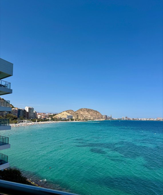 A scenic view of a turquoise sea meeting a sandy beach, with mountains and buildings in the distance under a clear blue sky.