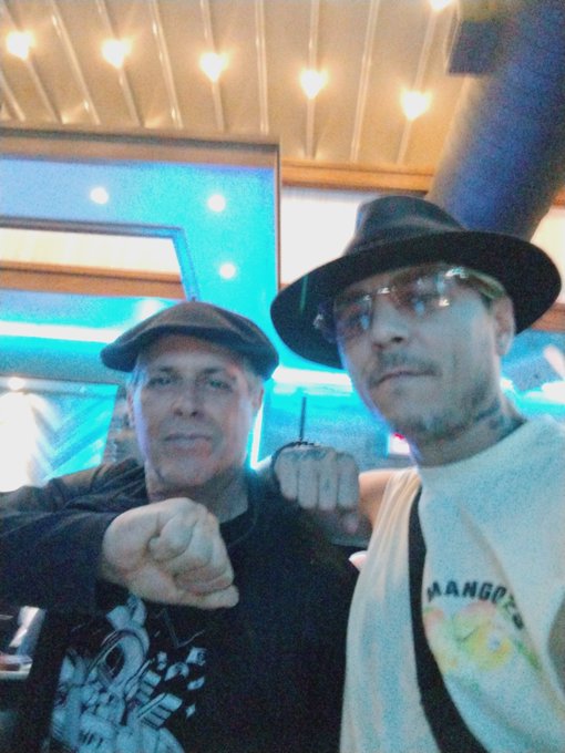 Two men pose for a selfie at an NFT NYC event, with one wearing a flat cap and graphic t-shirt making a fist gesture, while the other sports a black fedora, sunglasses, and a "BANGOR" tank top. The post tags @cybotcam and @NFT_NYC, suggesting the photo captures a friendly meetup between crypto/NFT community members at the New York City conference.