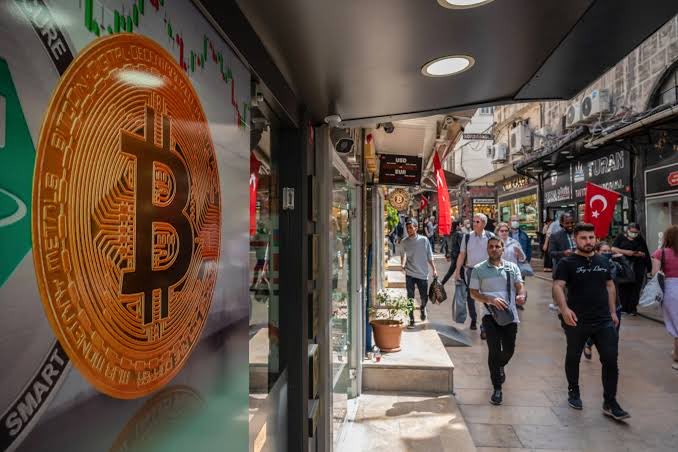 A storefront window displays a large orange Bitcoin logo with "DIGITAL DECENTRALIZED PEER TO PEER" and "SMART" text, while pedestrians walk down a busy Turkish street where a Turkish flag is visible in the background, illustrating the post about the massive financial losses suffered by Turkish crypto investors.