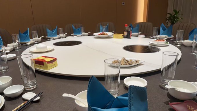 A pristine private dining room awaits its guests, featuring a large circular table set with individual cooking stations, neatly folded blue napkins, glasses, and small dishes of appetizers. A visible "LUCKY CARD" adds a playful touch to the anticipatory atmosphere, perfectly capturing the quiet excitement before friends arrive to share the meal.