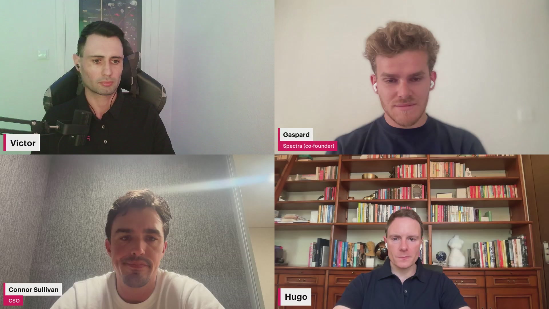 The image shows four men sitting at a table for a video call using a streaming platform like Zoom or Skype. The men are all wearing headphones and appear to be engaged in a conversation with each other. They are positioned around the table, facing one another as they communicate over the video call.