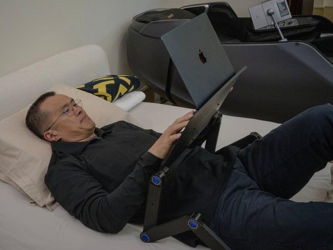 A man with glasses lying on a couch using a tablet mounted on a stand, with a MacBook laptop resting nearby on the couch.