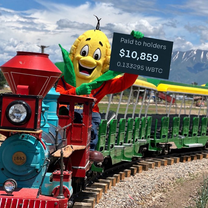 A person in a corn mascot costume riding a small red and green train while holding a sign that reads "Paid to holders $10,859 0.133295 WBTC".