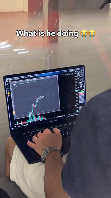 A person sitting and using a laptop displaying a stock/crypto trading chart with green and red candles, captioned with "What is he doing" and crying-laughing emojis.