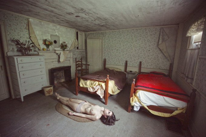 A person lies on the floor of a weathered, dilapidated bedroom, an image that visually represents the heavy theme of "The Weight Of Everything Is Unbearable." This portrait is a 1/1 piece available on @objktcom at objkt.com/tokens/KT1GQDs.