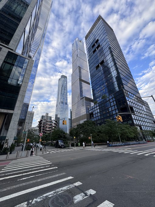 A modern Manhattan streetscape features towering glass skyscrapers, including what appears to be One57, alongside a wide intersection with crosswalks and traffic lights under a partly cloudy sky, capturing the urban backdrop as attendees head to the Avalanche Summit.