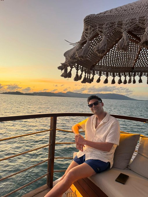 A man wearing sunglasses, a white short-sleeve shirt, and dark shorts sits on a cushioned boat bench with a phone beside him, posing with a sunset over the ocean and distant mountains in the background.