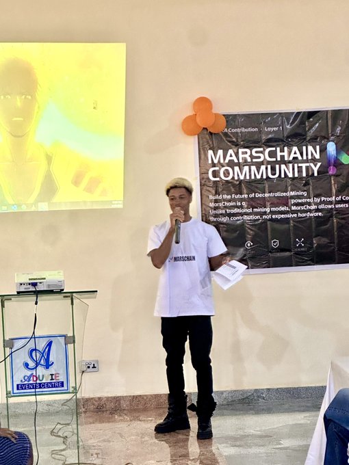 A young man in a white MarsChain t-shirt speaks into a microphone on stage, standing in front of a black "MarsChain Community" banner that promotes "Decentralized Mining" and "Proof of Contribution," with an adjacent screen displaying "Layer 1" against a yellow backdrop. The image visually captures the speaker anchoring the MarsChain web3 event referenced in the post, with the "SAU DIE Events Centre" podium in the foreground tying the scene to the venue where the gathering took place.