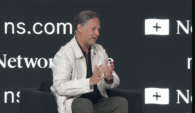 A man with gray hair wearing a light-colored jacket sits on a stage, gesturing with his hands as he speaks at what appears to be a conference with logos in the background.