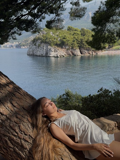 A woman with long hair rests peacefully against a tree trunk by the sea in a scenic coastal setting.