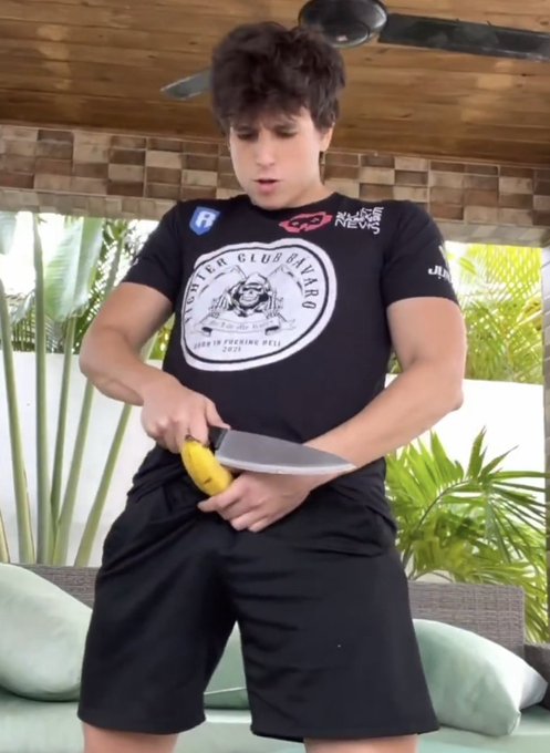 A young man with curly hair wearing a black 'Ninh Club Bistro' t-shirt stands outdoors under a wooden ceiling, peeling a banana with a large knife.
