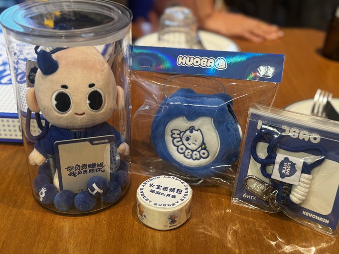 A collection of HUOGU branded merchandise, including a plush doll in a clear container, a small plush in packaging, a keychain, and tape, displayed on a wooden table with Chinese text visible on the doll's card.