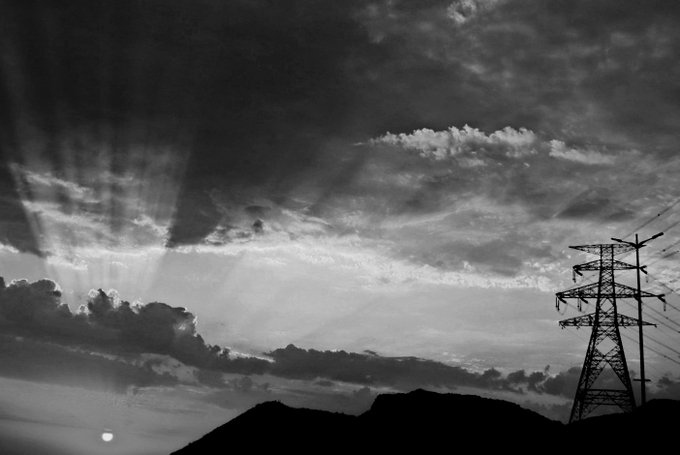 A black-and-white photograph captures dramatic crepuscular rays streaming down from behind clouds over a silhouetted landscape, with an electrical transmission tower on the right and the sun low on the horizon.