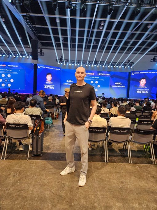 A bald man wearing a black "CryptoJobsList" t-shirt stands smiling in the foreground of a packed conference hall, with "AMBCZECH ASTRA," "METAPLANET," and "0.85" visible on the stage screens behind him, capturing his on-the-ground presence at BitcoinConfAsia in Hong Kong.