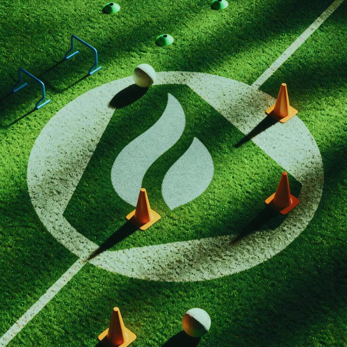 An overhead view of a soccer training field featuring a large flame logo painted on the grass, surrounded by orange traffic cones, white soccer balls, and small blue training hurdles.