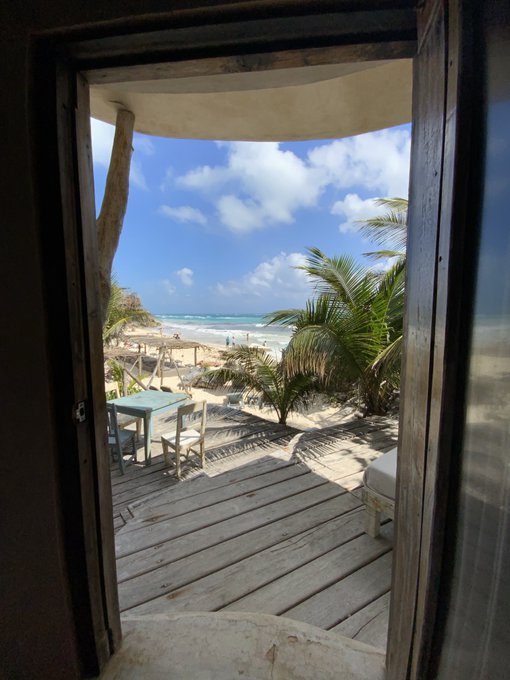 A view through an open wooden doorway reveals a rustic outdoor patio with a table and chairs, overlooking a sandy beach and ocean with gentle waves under a blue sky with scattered clouds.
