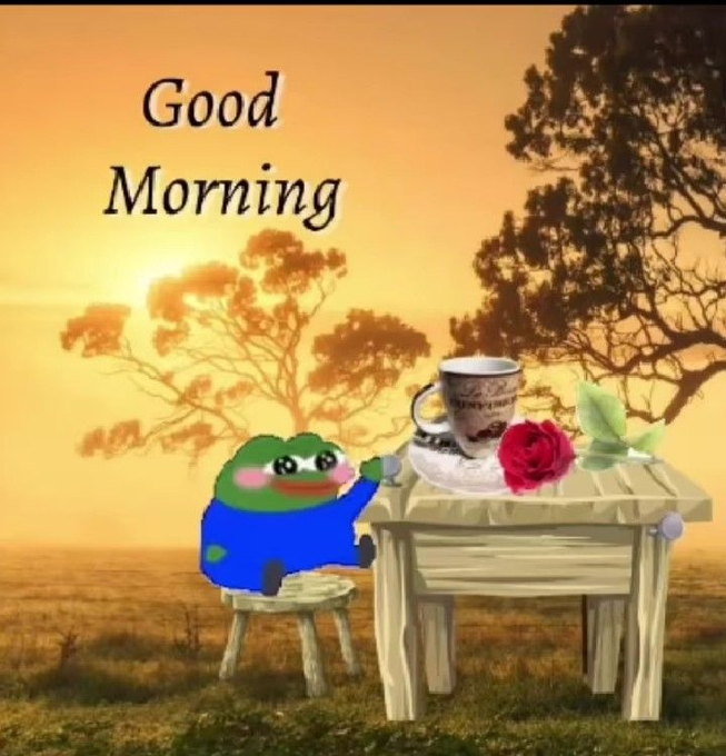 A 'Good Morning' greeting image featuring a small cartoon frog sitting at a wooden table with a coffee cup and a red rose against a sunset landscape.