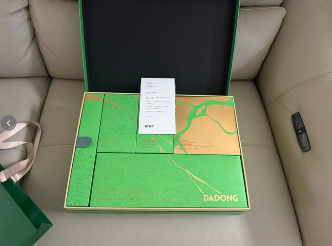 The image shows a green box with gold lettering that reads "DADONG", placed on top of a couch cushion. The box appears to be open, possibly containing a gift or item for sale.