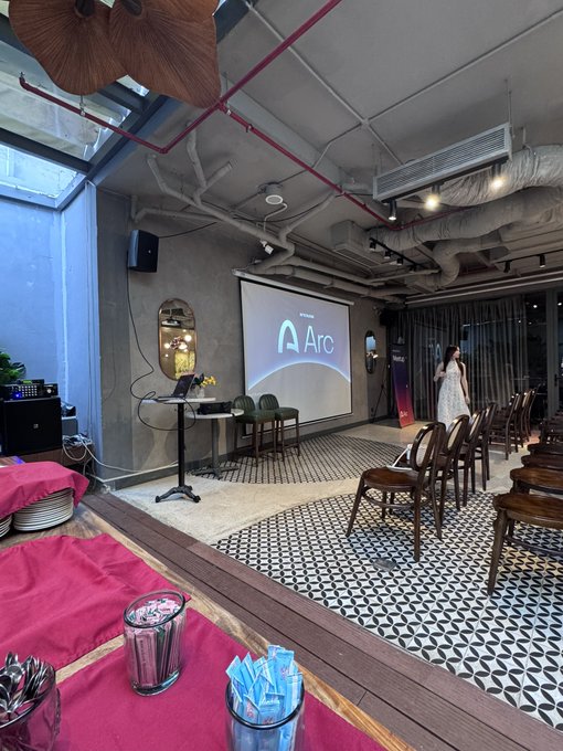 An indoor event space with exposed ceiling pipes, patterned tile floors, and rows of wooden chairs facing a projection screen displaying the logo 'Aro'.