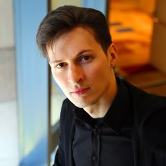 A portrait photograph of a young man with short dark hair wearing a black shirt, standing near a window.