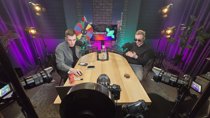 Two men sit across from each other at a wooden table in a studio setup with professional cameras, lighting, and a colorful wheel in the background.